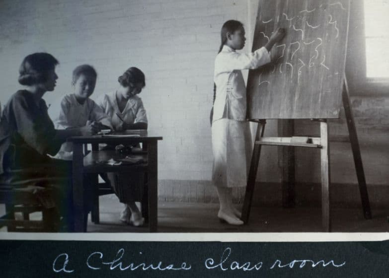 Black and white image depicting a teacher writing on a blackboard in front of three students, with the caption, "A Chinese classroom."